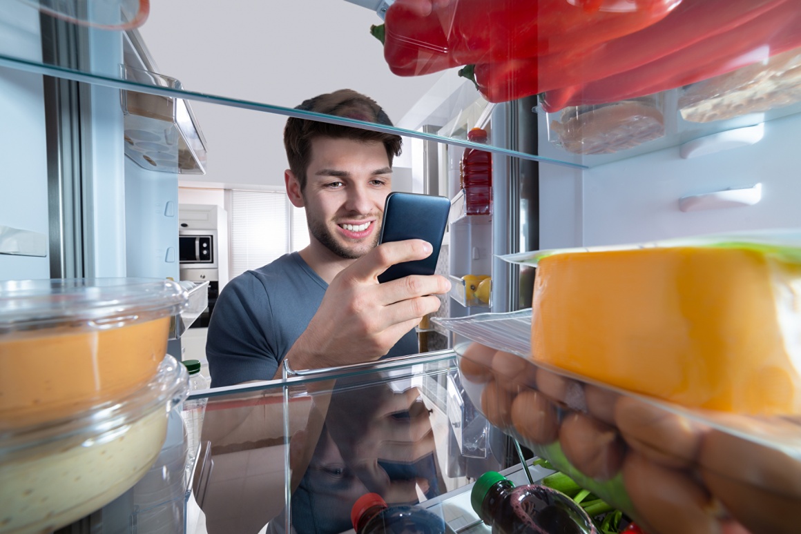 A young man looking into his fridge and holding a smartphone...