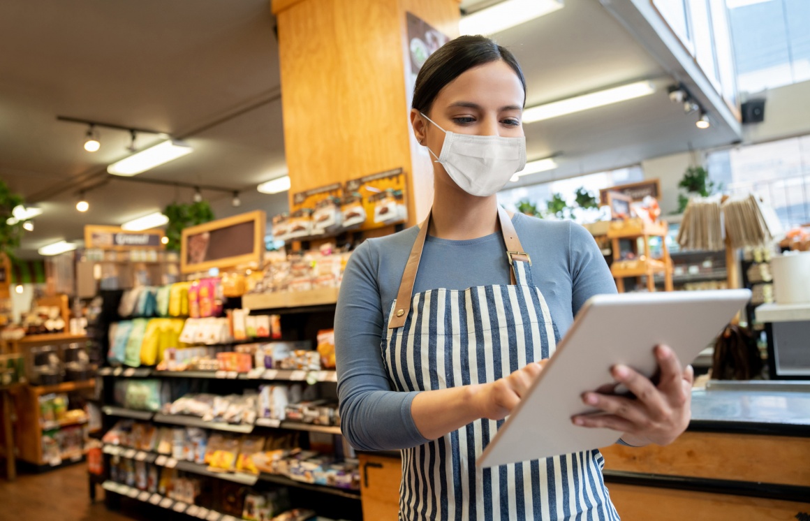 A saleswoman with an apron and face mask holds a Tablet PC in her hand...