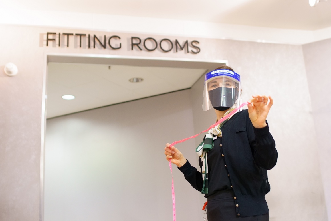 A woman with face mask and measuring tape in front of a fitting room...