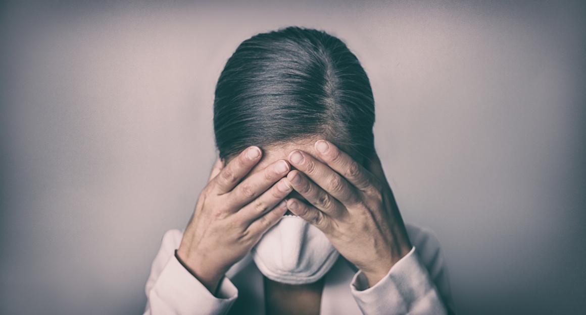 A woman with a face mask on is hiding her face in her hands...