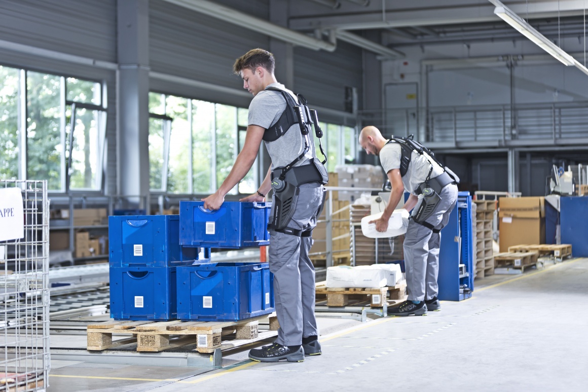 Two men lift boxes wearing exoskeletons