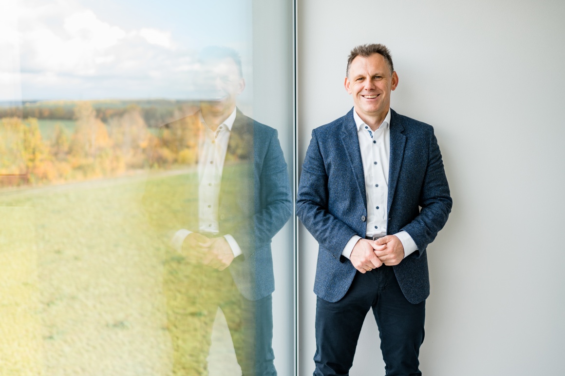 Man in suit looks into camera and stands next to window through which landscape...