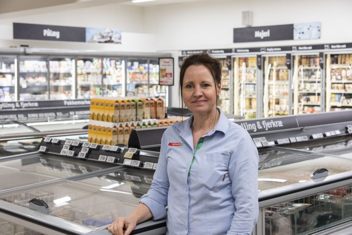 Smiling woman stands in grocery store next to a freezer compartment...