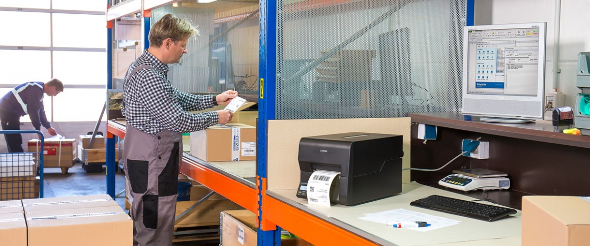Two shipping employees stand in a hall next to a shipping label printer and...